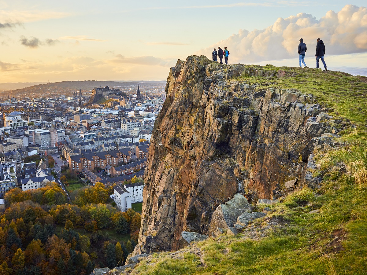 Group of people walking along cliff edge looking at city views. Edinburgh Castle in the distance